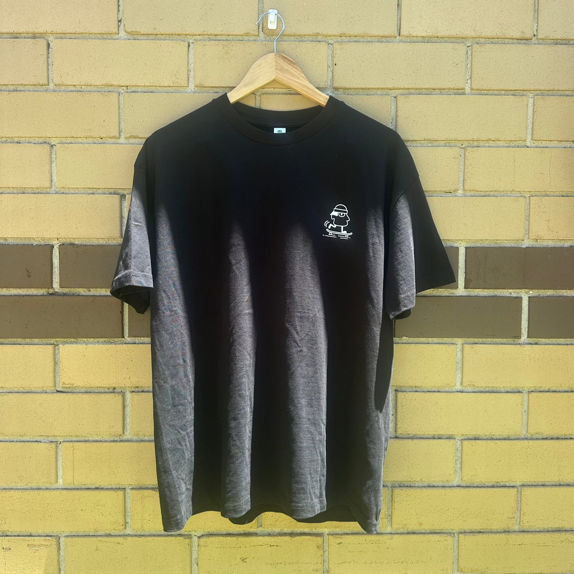 Black t-shirt with a small white logo on a hanger against a brick wall.